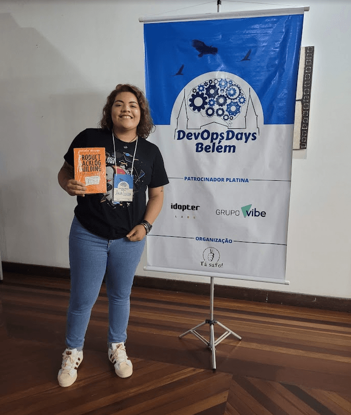 Ana Luiza Primo ao lado do banner do DevOps Days Belém, segurando um livro